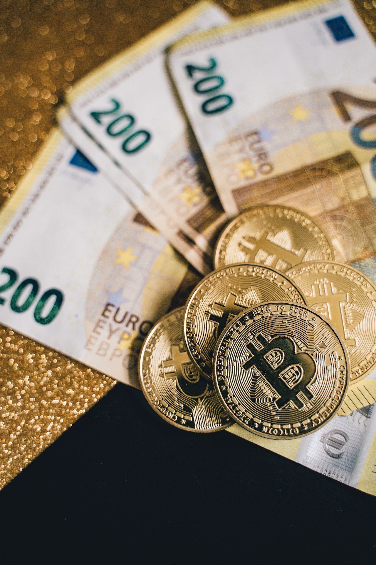 Euro banknotes (€200) and gold cryptocurrency coins on a glittery surface, showing cash and Bitcoin-like symbols without context of use