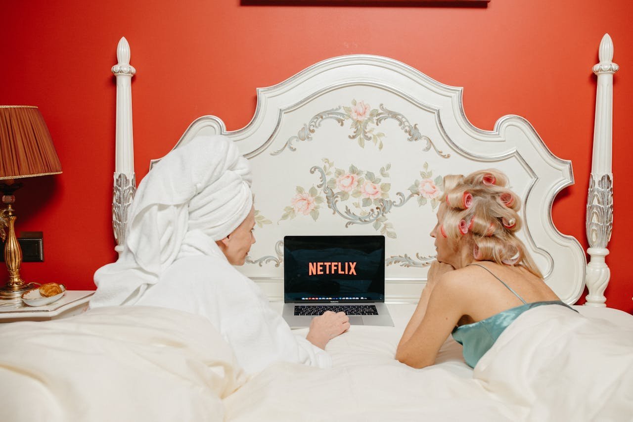 Couple in bed watching Netflix on a laptop, one with a towel turban and the other with pink curlers.