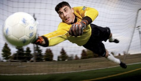 Soccer goalkeeper in a yellow jersey dives to block a white ball near the goal, dramatic action mid-air.