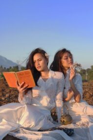 Two women in white dresses reading a book together on a blanket outdoors, with mountains in the background.