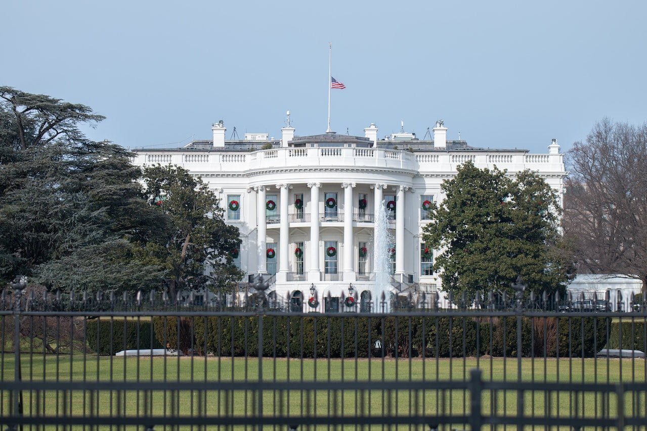Front view of the White House with holiday wreaths on the columns, an American flag flying, seen behind a black metal fence.