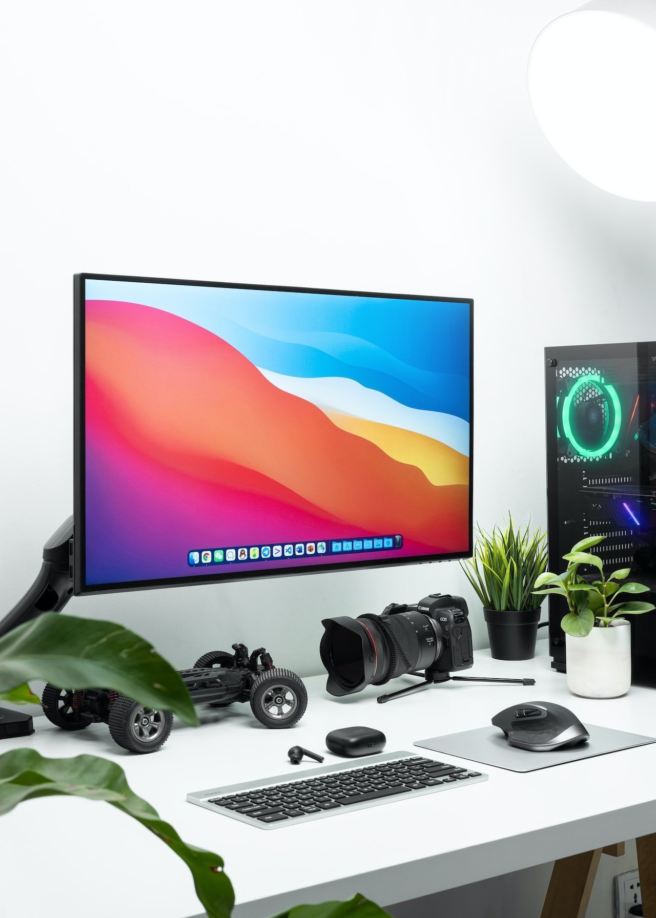 Desk setup with a large monitor showing colorful wallpaper, surrounded by camera gear, keyboard, mouse, and plants on a white desk.