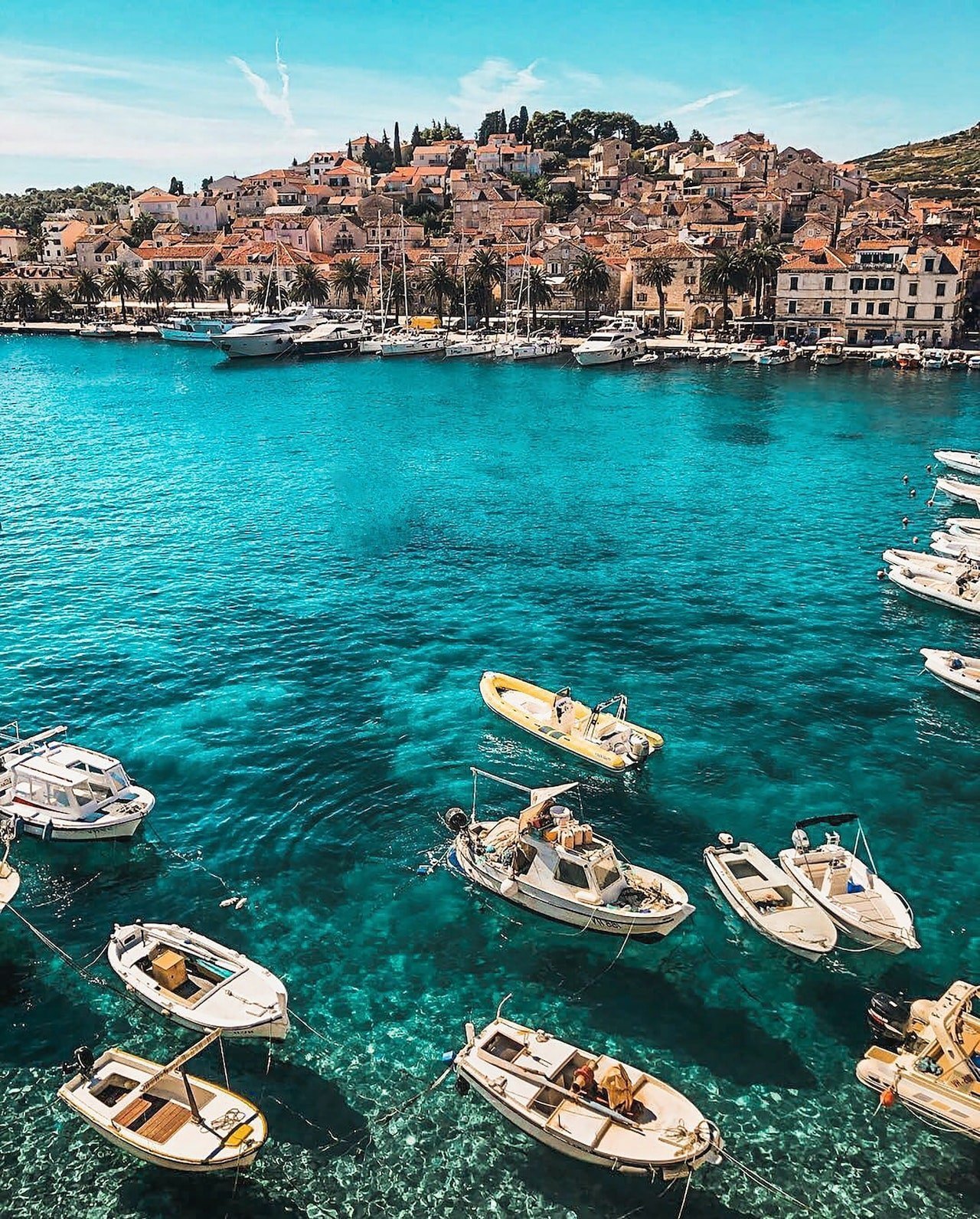 Harbor scene in a Mediterranean town: turquoise water with small boats anchored near a rocky, sunlit shoreline and a hillside of stone houses behind palm trees