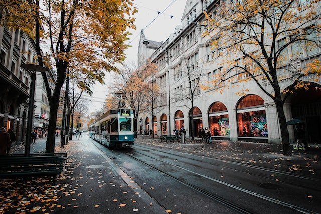 Tram on a wet city street in autumn, with shop archways and fallen orange leaves nearby.