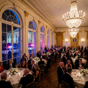 Formal dinner with guests seated at round tables in White House room with chandeliers and police cars visible outside windows