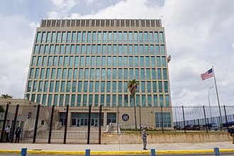 Modern glass-front office building behind a black fence, with a palm tree and an American flag in front on a cloudy day.