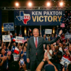Male politician on stage at a Texas victory rally, crowd waving flags and signs like 'Texas Votes' and 'We Won!' behind him under a large 'Victory' banner with 'KEN PAXTON' text (avoid naming the person).