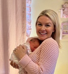 Smiling woman in a pink sweater holds a newborn baby wrapped in a white blanket in a pastel nursery.