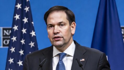 Man in a dark suit and blue tie speaks at a podium, microphones in front, with the American flag and a blue backdrop behind him.