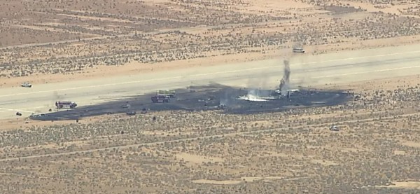 Desert scene of a burned aircraft wreck on or beside a road, with black smoke rising and several emergency or support vehicles nearby.