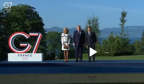 Three people in formal attire stand on a stage next to a large G7 France Evian sign, with a blue sky and mountainous landscape in the background.